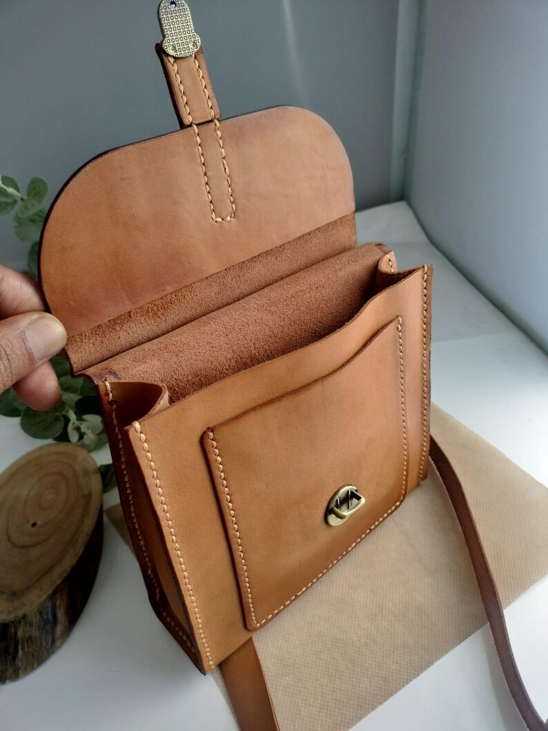 May include: A light brown leather crossbody bag with a flap closure and a front pocket. The bag has a long, adjustable strap and a metal clasp. The stitching is visible and the bag appears to be handmade.