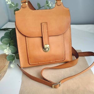 May include: A light brown leather crossbody bag with a flap closure and adjustable strap. The bag features a rectangular design with a rounded flap, brass-colored hardware, and visible stitching. The bag is approximately 9 inches tall and 8 inches wide.