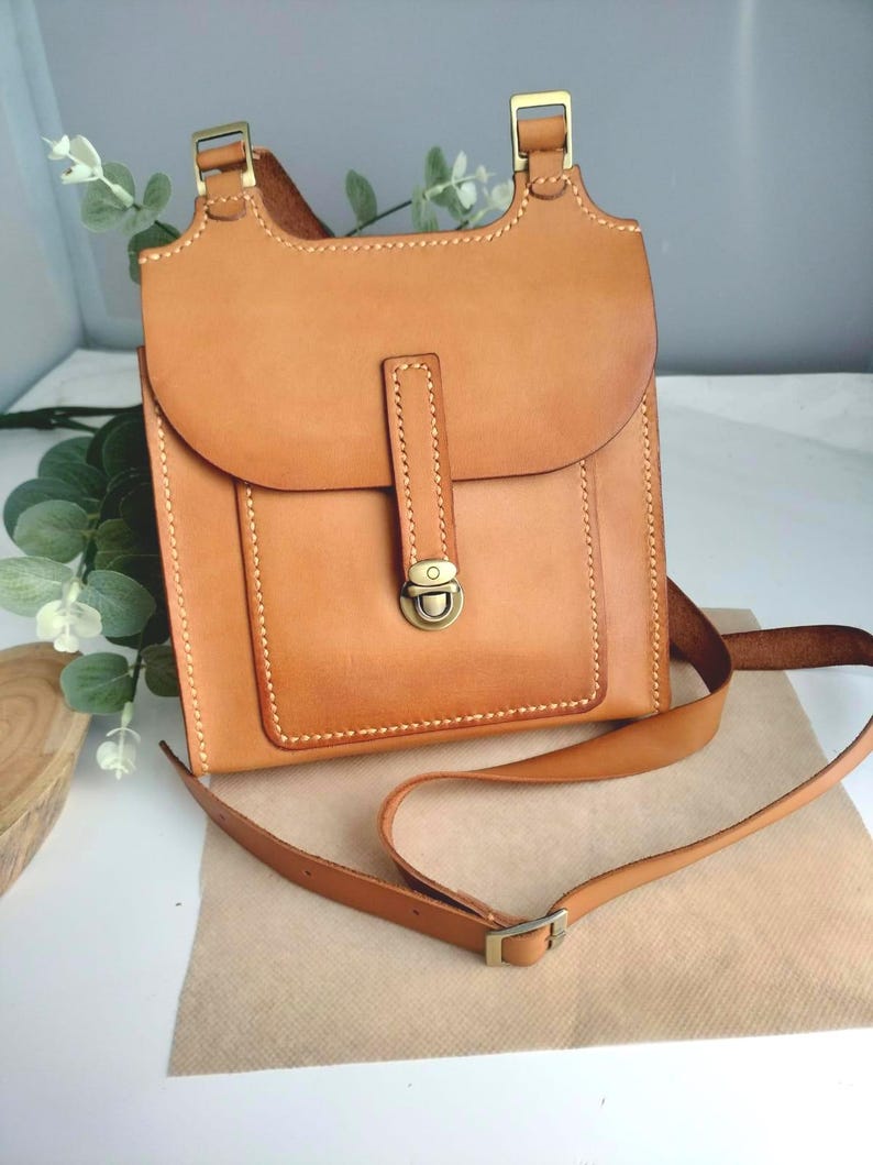May include: A light brown leather crossbody bag with a flap closure and adjustable strap. The bag features a rectangular design with a rounded flap, brass-colored hardware, and visible stitching. The bag is approximately 9 inches tall and 8 inches wide.