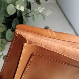 May include: Close-up of a handcrafted, light brown leather bag with visible stitching and a metal clasp. The bag's interior is a lighter shade of brown, and the design suggests a structured, possibly vintage style. The background includes greenery.