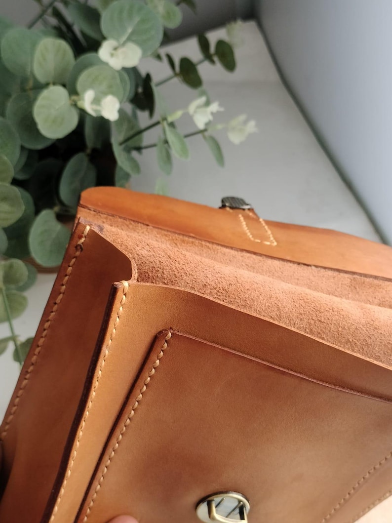 May include: Close-up of a handcrafted, light brown leather bag with visible stitching and a metal clasp. The bag's interior is a lighter shade of brown, and the design suggests a structured, possibly vintage style. The background includes greenery.