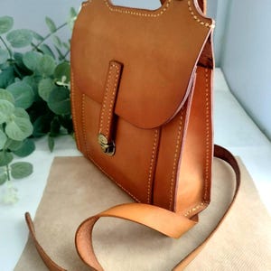 May include: A handcrafted, medium-sized, brown leather bag with a flap closure and a shoulder strap. The bag features visible stitching and a metal clasp. The bag has a top handle and a long adjustable strap. The leather has a smooth, polished finish.