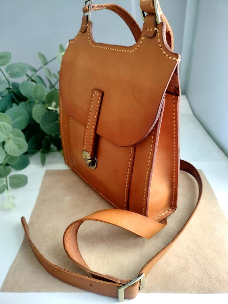May include: A handcrafted, medium-sized, brown leather bag with a flap closure and a shoulder strap. The bag features visible stitching and a metal clasp. The bag has a top handle and a long adjustable strap. The leather has a smooth, polished finish.