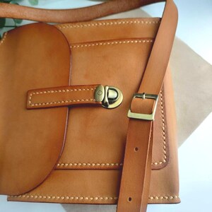 May include: A handcrafted, light brown leather crossbody bag with a flap closure and adjustable strap. The bag features a brass buckle and stitching details. The bag is designed with a rectangular shape and a rounded flap.
