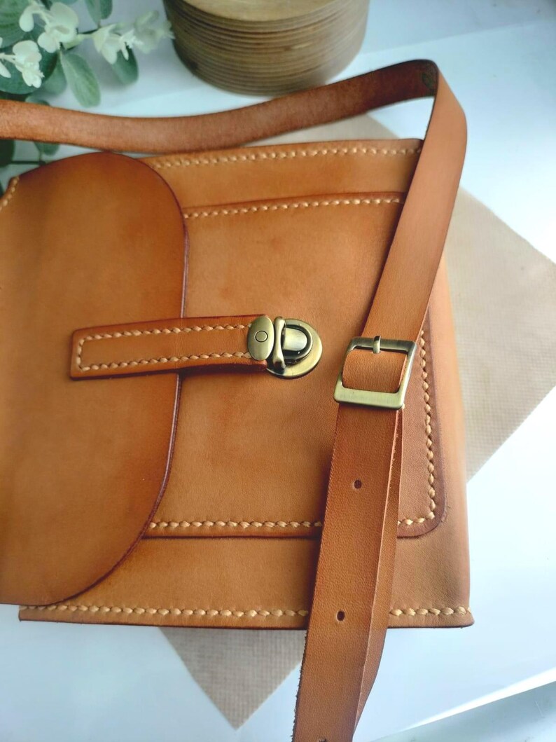 May include: A handcrafted, light brown leather crossbody bag with a flap closure and adjustable strap. The bag features a brass buckle and stitching details. The bag is designed with a rectangular shape and a rounded flap.