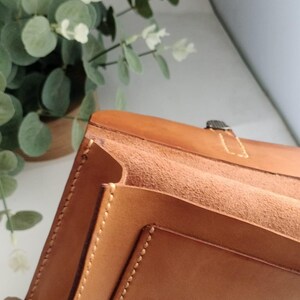 May include: A light brown leather portfolio with visible stitching and a metal clasp. The interior features multiple compartments. The portfolio is open, revealing the textured interior. The background includes greenery.