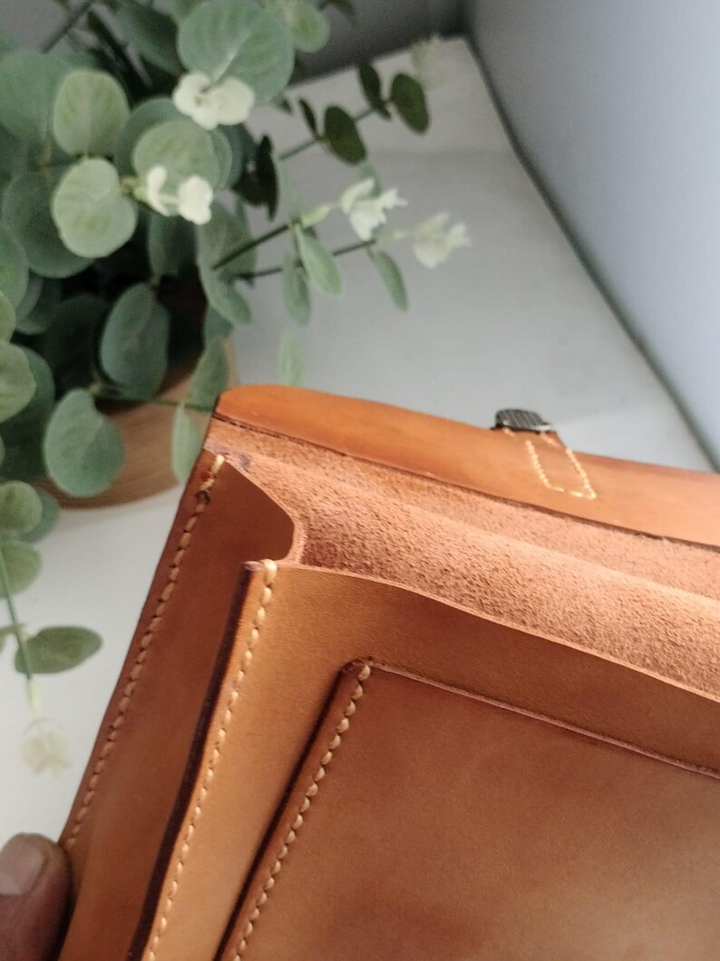 May include: A light brown leather portfolio with visible stitching and a metal clasp. The interior features multiple compartments. The portfolio is open, revealing the textured interior. The background includes greenery.