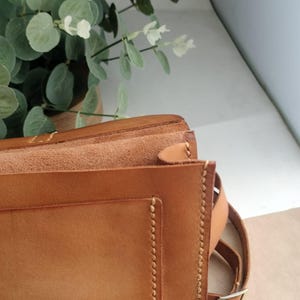 May include: A close-up of a tan leather satchel bag with a flap closure. The bag features visible stitching along the edges and a brown adjustable strap. The background includes green and white foliage.