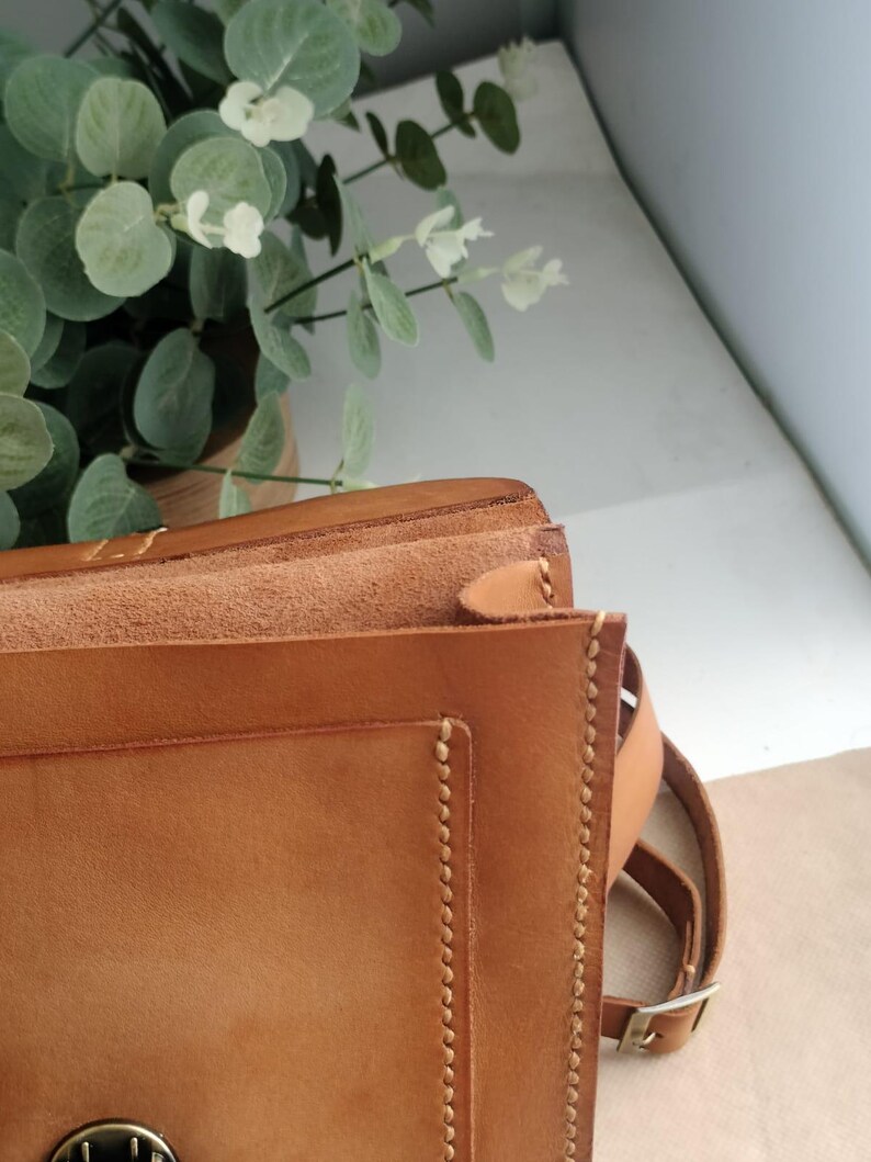 May include: A close-up of a tan leather satchel bag with a flap closure. The bag features visible stitching along the edges and a brown adjustable strap. The background includes green and white foliage.
