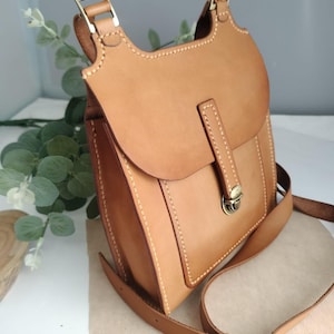 May include: A light brown leather crossbody bag with a flap closure and adjustable strap. The bag features brass-colored hardware and visible stitching. The bag is designed with a rectangular shape and a rounded top.