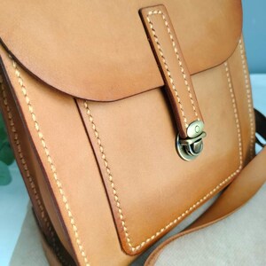 May include: A tan leather crossbody bag with a flap closure and a shoulder strap. The bag features visible stitching and a metal clasp. The leather has a smooth texture and a natural color, suggesting a handcrafted design. The bag is designed for carrying essentials.
