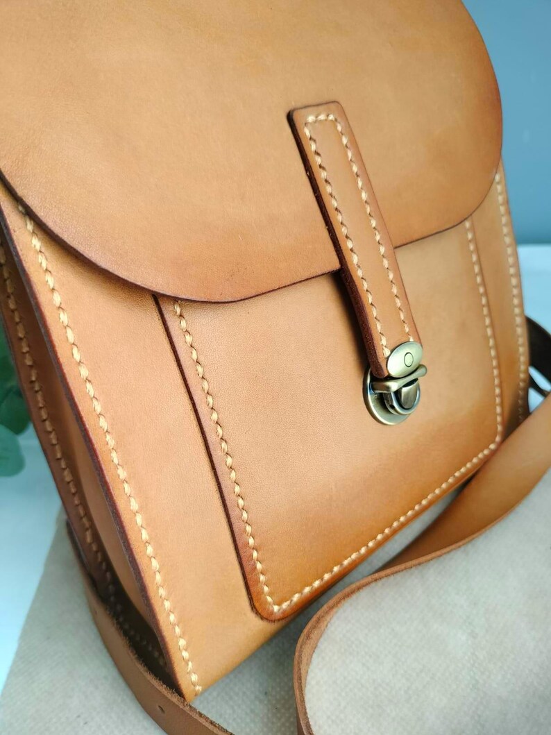 May include: A tan leather crossbody bag with a flap closure and a shoulder strap. The bag features visible stitching and a metal clasp. The leather has a smooth texture and a natural color, suggesting a handcrafted design. The bag is designed for carrying essentials.