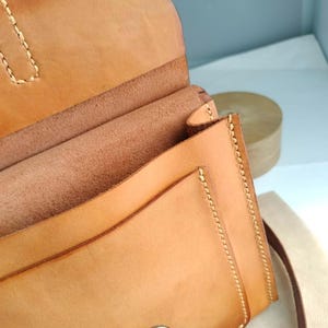 May include: A light brown leather crossbody bag with a flap closure and a metal clasp. The bag features visible stitching along the edges and a long strap. The interior of the bag is visible, showing multiple compartments.