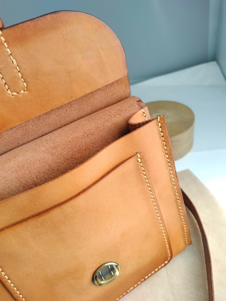 May include: A light brown leather crossbody bag with a flap closure and a metal clasp. The bag features visible stitching along the edges and a long strap. The interior of the bag is visible, showing multiple compartments.