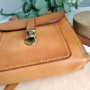 May include: A tan leather crossbody bag with a flap closure and a metal clasp. The bag features visible stitching and a long strap. The bag is made of a light brown leather and has a rectangular shape. The bag is a handmade item.