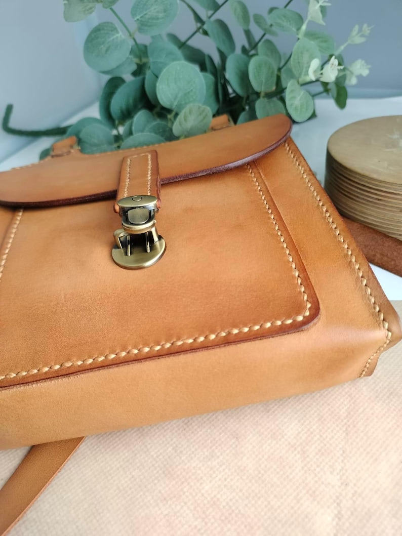 May include: A tan leather crossbody bag with a flap closure and a metal clasp. The bag features visible stitching and a long strap. The bag is made of a light brown leather and has a rectangular shape. The bag is a handmade item.