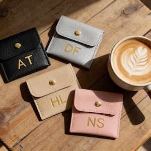 Könnte beinhalten: Draufsicht auf vier kleine, personalisierte Leder-Münzbörsen in Schwarz, Grau, Beige und Rosa, jeweils mit goldenen Buchstaben und einem goldenen Druckknopfverschluss. Rechts steht eine Tasse Latte Macchiato mit Latte Art auf einer Holzoberfläche.