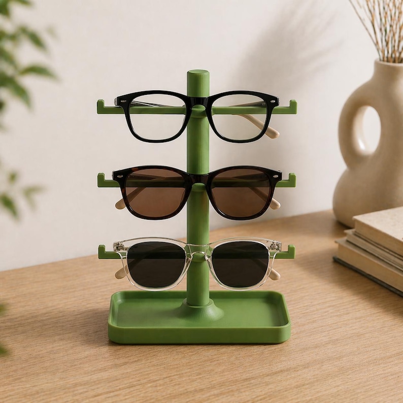 May include: A green sunglasses display stand holding three pairs of sunglasses. The sunglasses have black, brown, and clear frames. The stand is made of metal and has a rectangular base.