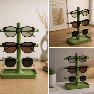 May include: A green sunglasses display stand holding three pairs of sunglasses. The sunglasses have black, brown, and clear frames. The stand is made of green plastic and has a small tray at the base.