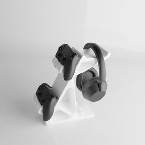 May include: A white gaming accessory stand holding two black game controllers and a pair of black headphones. The stand is designed to organize gaming equipment and has a modern, minimalist aesthetic.
