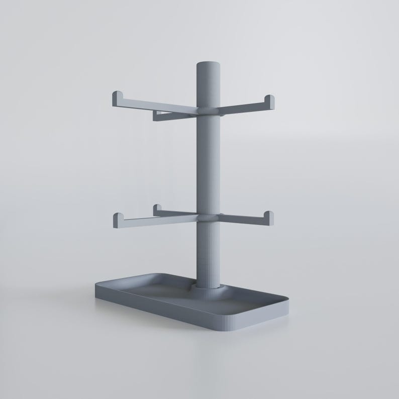 May include: A gray, tiered mug rack with a rectangular base and three levels of hooks. The central pole supports the horizontal arms, designed to hold mugs. The rack is made of a solid, matte material.