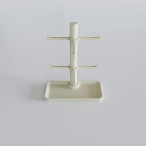 May include: A cream-colored mug tree with a rectangular base and a central pole. The pole has two tiers of hooks for hanging mugs, and a cylindrical top. The design is minimalist and functional, suitable for kitchen organization.