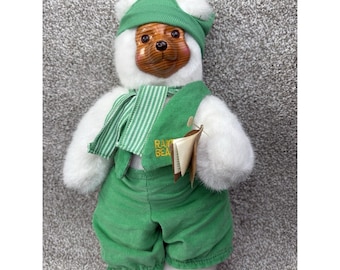 Könnte beinhalten: Ein Plüschteddybär mit grünem Hut, Latzhose und Schal. Der Bär hat ein holzähnliches Gesicht und weißes Fell. Auf der Latzhose steht der Text "RAW BEA". Ein Etikett ist an der Latzhose des Bären befestigt.
