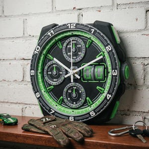 May include: A large black and green wall clock designed to resemble a wristwatch. The clock features multiple subdials, white hands, and a digital display. A green toy car, gloves, and keys are on a wooden surface in front of a white brick wall.