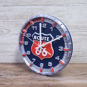 Op de afbeelding: Een ronde wandklok met een Route 66-ontwerp. De klok heeft een marineblauwe wijzerplaat met witte cijfers en wijzers. Het Route 66-logo is rood en wit. De klok heeft een zilveren en rode rand. De diameter is ongeveer 20 cm.