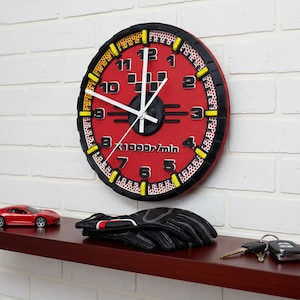 May include: A red and black wall clock with white hands, resembling a car's speedometer, is mounted on a white brick wall. A red toy car, black gloves, and keys rest on a dark brown shelf below the clock.