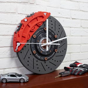 Op de afbeelding: Een wandklok ontworpen als een remschijf van een auto met een rode remklauw. De klok heeft witte wijzers en is gemonteerd op een witte bakstenen muur. Een zilveren speelgoedauto, autosleutels en racehandschoenen liggen op een houten oppervlak.