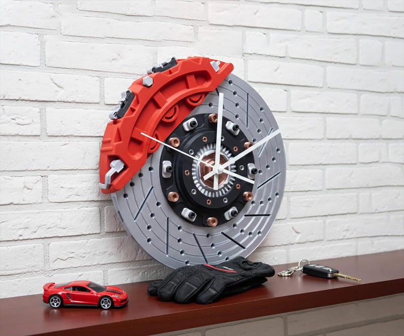 Car brake disc styled wall clock. Red calipers and silver disc. White hour, minute and second hands. Black center hub with bronze bolts. On a wooden shelf, leaned against a white brick wall, with a racing car model, gloves and keys. Sim racing car decor perfect for car gifts for men and garage sign.
