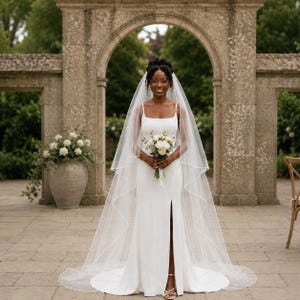 May include: A floor-length white wedding dress with a square neckline, thin straps, and a front leg slit. The dress is paired with a long, sheer, floor-sweeping veil with a delicate piped edge. A bouquet of white roses and greenery is held in front.