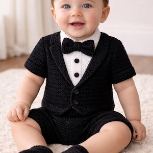 Puede incluir: Un conjunto de esmoquin de bebé de ganchillo negro, que incluye una chaqueta con cuello blanco, pantalones cortos, una pajarita y botines. El atuendo tiene una parte delantera abotonada y lo usa un bebé sentado en una alfombra blanca.