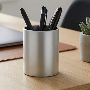 Puede incluir: Un portalápices cilíndrico plateado lleno de bolígrafos negros está sobre un escritorio de roble claro. Una computadora portátil cerrada, una taza y una pequeña suculenta en una maceta de concreto también están en el escritorio.