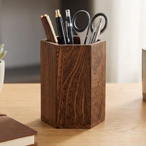 Könnte beinhalten: Ein sechseckiger Schreibtisch-Organizer aus dunkelbraunem Holz. Er enthält Stifte, Bleistifte und eine Schere. Eine kleine Sukkulente im Topf, eine Tasse und ein ledergebundenes Notizbuch befinden sich ebenfalls auf dem Schreibtisch.
