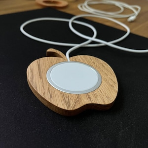 Könnte beinhalten: Drahtloses Ladegerät in Apfelform aus hellem Holz mit weißer Ladefläche und silbernem Rand. Das Ladegerät hat ein weißes Kabel. Es liegt auf einer schwarzen Oberfläche.