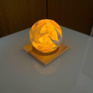 以下が含まれることがあります： 葉の模様が施された、光る球形のランプ。正方形の木製の台座の上に置かれています。ランプは温かみのある黄色い光を放ち、居心地の良い雰囲気を作り出しています。台座は明るい色の木材で作られています。装飾品です。