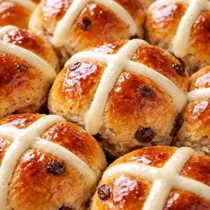 May include: Close-up of several golden-brown hot cross buns. Each bun has a white cross on top and is dotted with dark raisins. The buns have a shiny glaze, suggesting they are freshly baked.