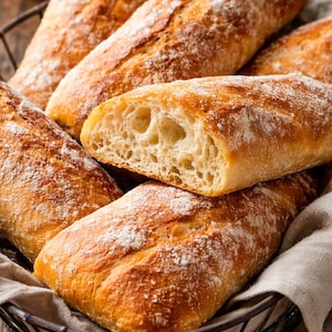 Op de afbeelding: Een mand gevuld met verschillende versgebakken broden. Het brood heeft een goudbruine korst, bestrooid met bloem, en een licht interieur. Eén brood is doorgesneden en toont de luchtige textuur.