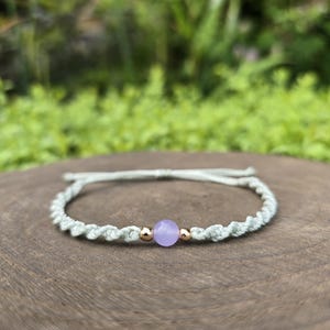 Könnte beinhalten: Ein hellgrünes Makramee-Armband mit einer einzelnen lavendelfarbenen Perle in der Mitte, die von zwei kleinen goldfarbenen Metallperlen flankiert wird. Die Kordel ist spiralförmig geflochten und hat verstellbare Zugbänder am Verschluss.