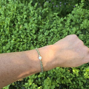 May include: A bracelet featuring a braided sage green cord with a single central white bead flanked by two small gold-toned metallic beads. The cord is woven in a tight, textured pattern, creating a simple and uniform band around the wrist.