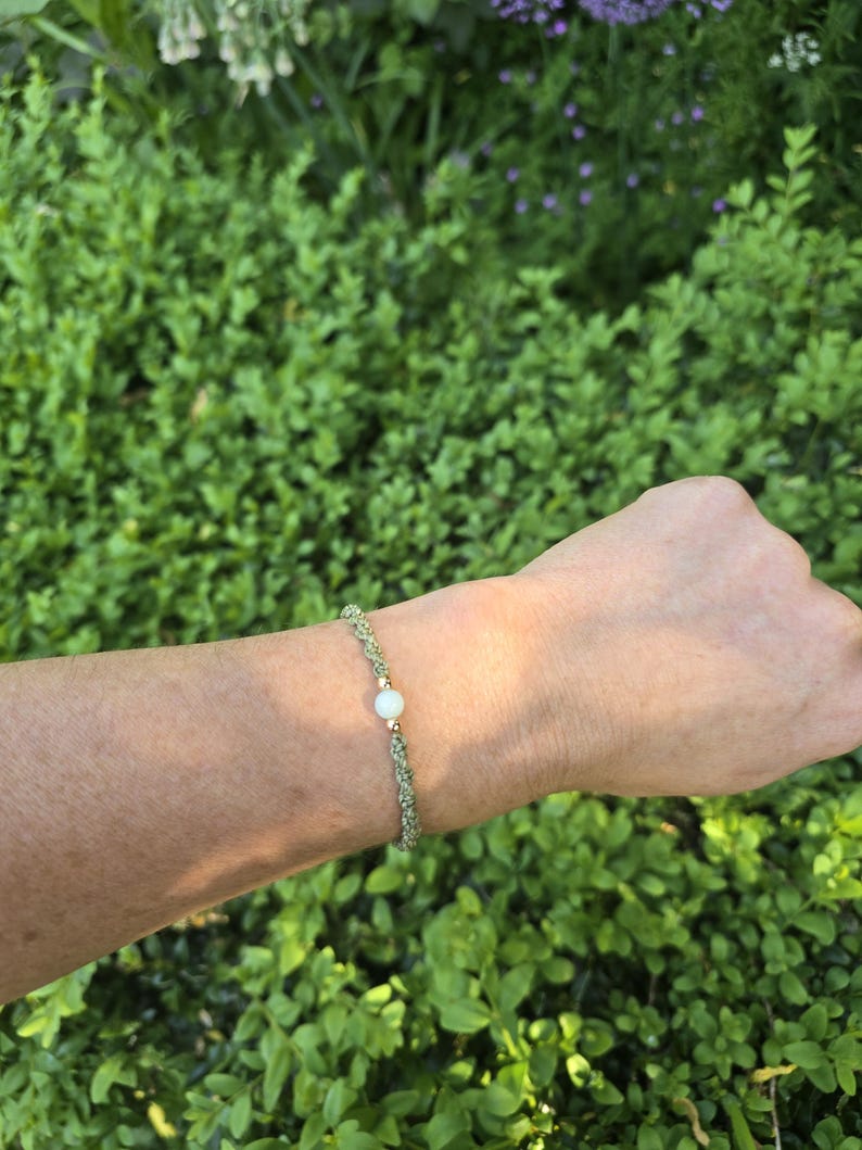 May include: A bracelet featuring a braided sage green cord with a single central white bead flanked by two small gold-toned metallic beads. The cord is woven in a tight, textured pattern, creating a simple and uniform band around the wrist.