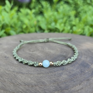 May include: A bracelet featuring a sage green cord with a braided design, centered with a single light blue round bead flanked by two small metallic gold-toned beads. The cord includes an adjustable sliding knot closure mechanism.