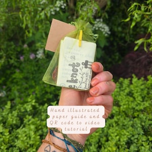 May include: A green organza drawstring pouch containing a hand-illustrated paper guide with the text "knot" and "pass the left knotting cord over". The pouch includes a small yellow clip and a brown cord, accompanied by three thin braided bracelets.