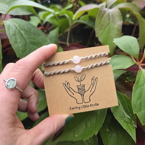 May include: Two beige twisted cord bracelets, each featuring a single pale pink round bead and small silver-toned accents. The bracelets are displayed on a rectangular tan card printed with a black line drawing of hands holding a plant and the text Earthy Little Knots.
