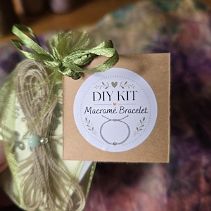 May include: A DIY kit for a macram&eacute; bracelet packaged in a sheer light green organza drawstring bag. A square brown cardboard tag is attached to the bag, featuring a white circular sticker with the text DIY KIT Macram&eacute; Bracelet and a graphic.