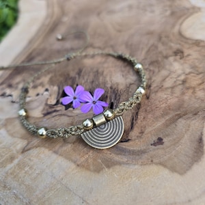 May include: A bracelet featuring a braided tan cord with metallic gold-toned beads and a central semi-circular charm embossed with concentric circles. The design includes adjustable cord ends and is accented by two small purple flowers.