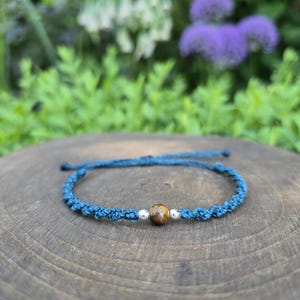 May include: A blue cord bracelet featuring a central brown tiger eye gemstone bead flanked by two small silver-toned metal beads. The bracelet is constructed with a knotted cord design and includes adjustable pull strings at the back for sizing.