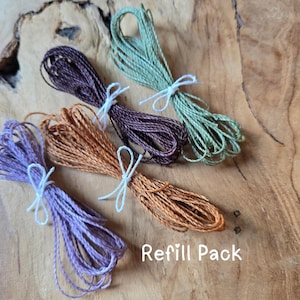 May include: Four bundles of twisted cord in light purple, orange, dark brown, and sage green, each tied with a thin white string. The text Refill Pack is printed in white at the bottom right. The cords have a textured, fibrous appearance.